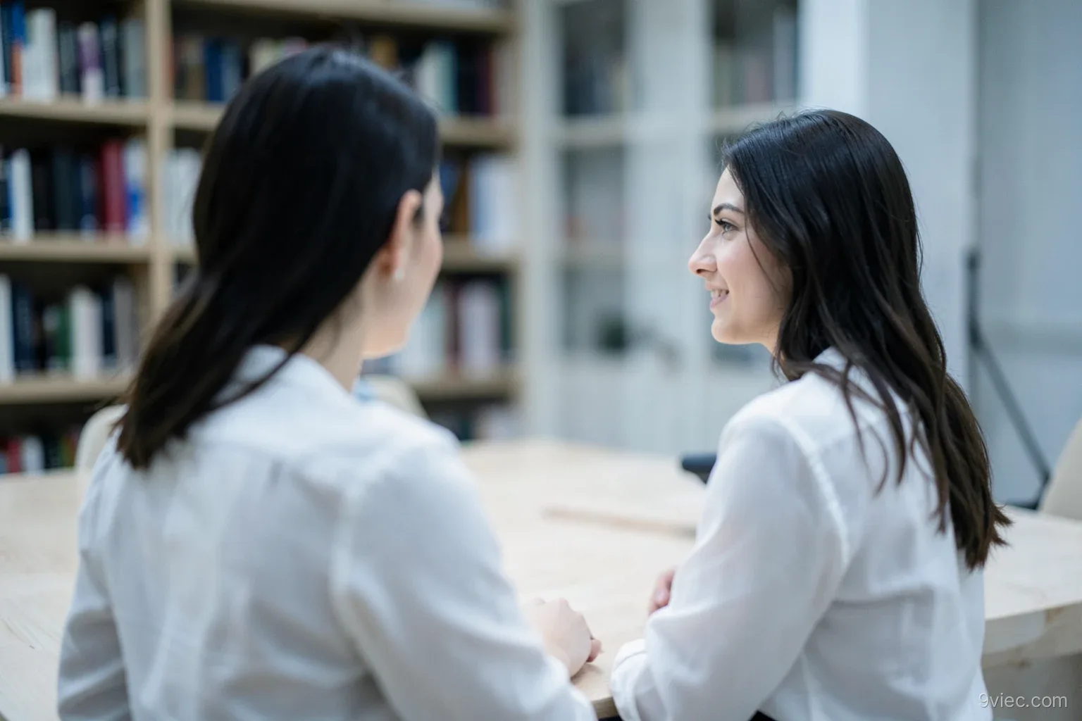Over-the-shoulder view of a young woman in a plain blouse listening to an interviewer across a long pale wooden table, blurred bookshelf behind, cool overcast light